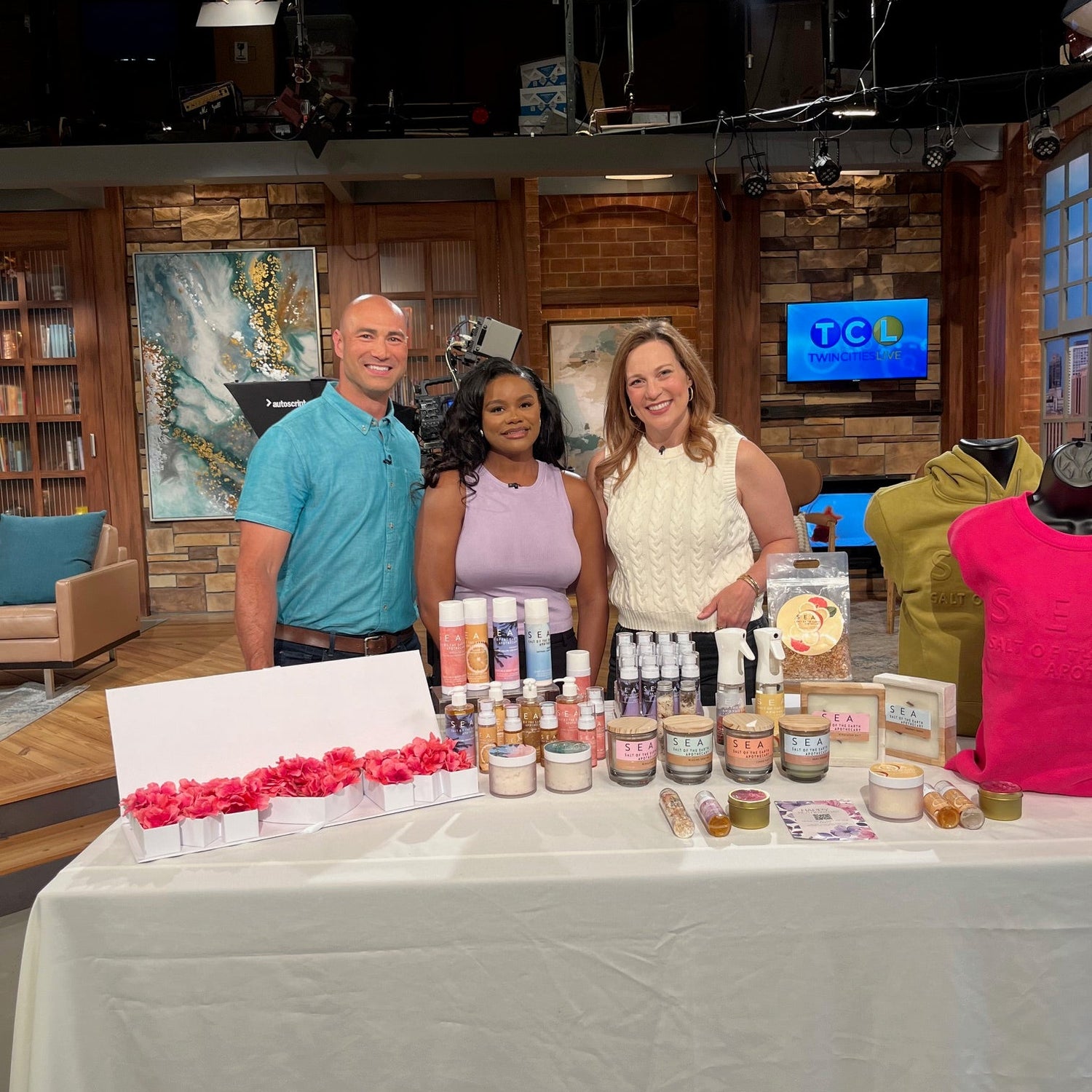 Two women and a man on the news standing behind a counter with various items on display, including a candle scrubs and oils .